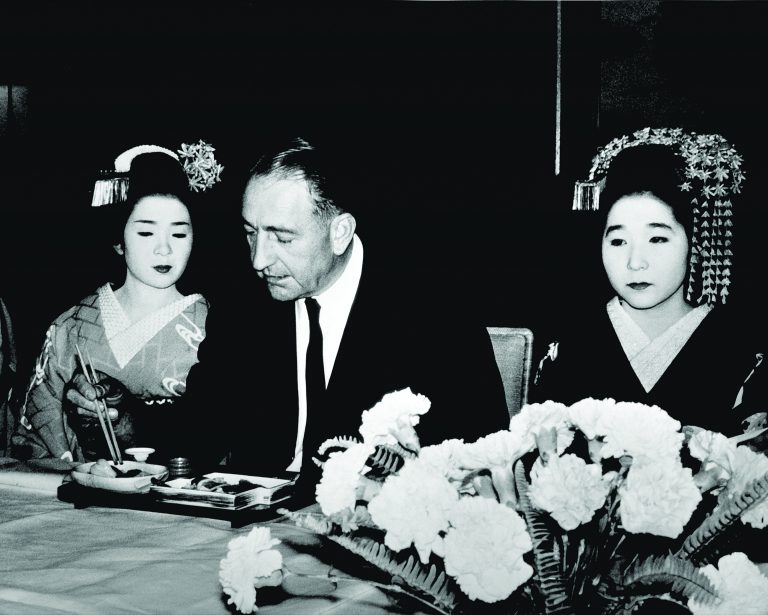 Dave Packard dining during his visit to Japan in 1963. Two women in traditional Japanese dress sit on either side of him.