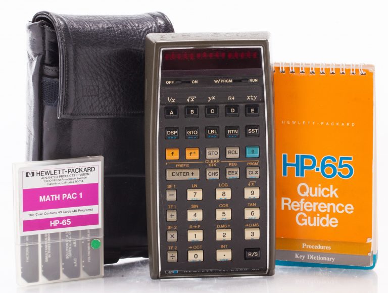 A photo of the HP 65 programmable pocket calculator with quick reference guide, a pack of inserts and carrying case.