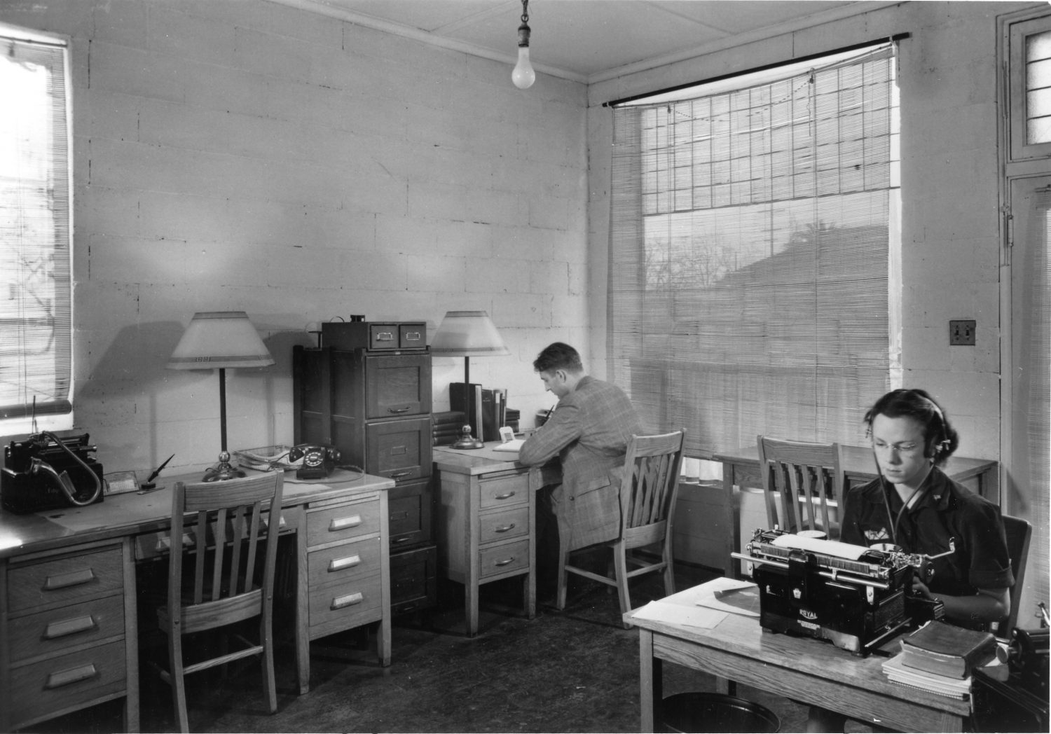 Photo of Helen Perry at work (typing with headset on) with Dave Packard working in the background.