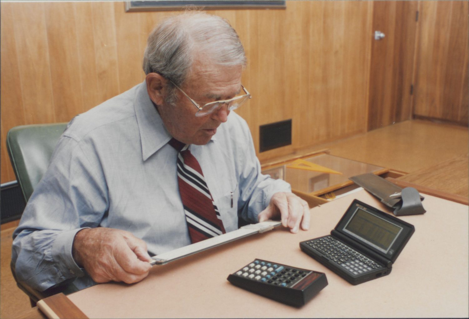 Bill Hewlett with a slide rule in his hand, while an HP 35 and HP 95LX palmtop lay on his desk.