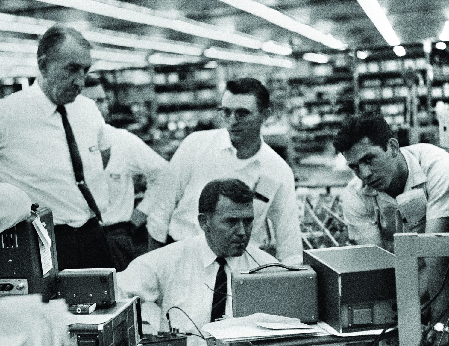 Bill Hewlett and Dave Packard interacting with employees. Hewlett is tinkering with an instrument while the others look on. 