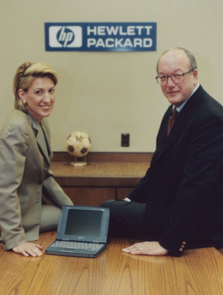 Carly Fiorina and Lew Platt seated on a table next to an HP laptop.